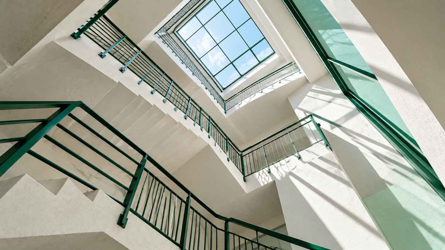 Upward view of a white staircase with green railings forming sharp diagonal lines and right angles under a bright skylight