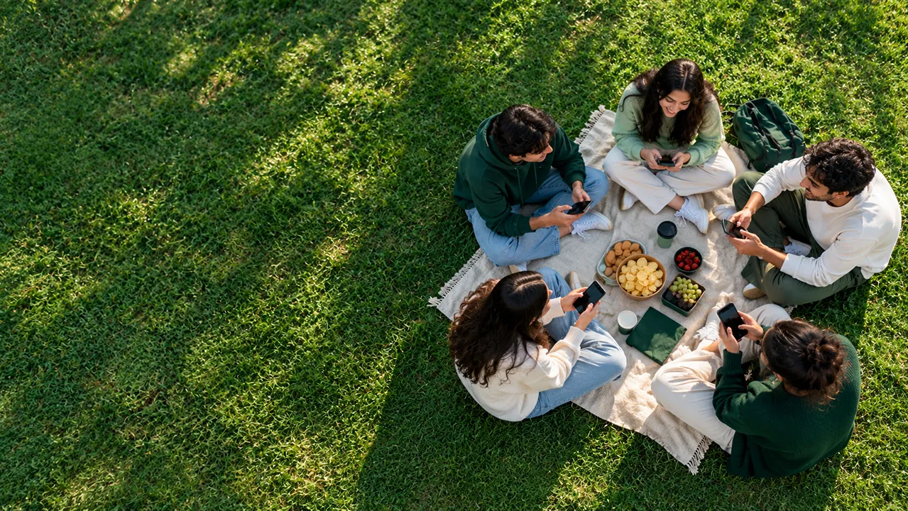 Aerial view of five friends sitting in a circle on a lawn, each looking at a phone